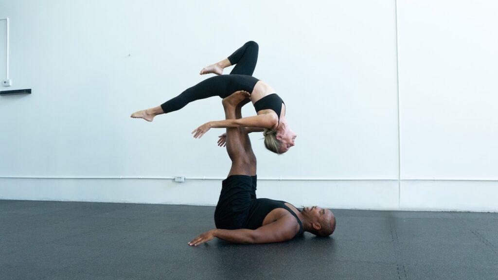 A man and woman doing partner acro. The man is on the floor with his legs in the air holding a woman in a back bend.